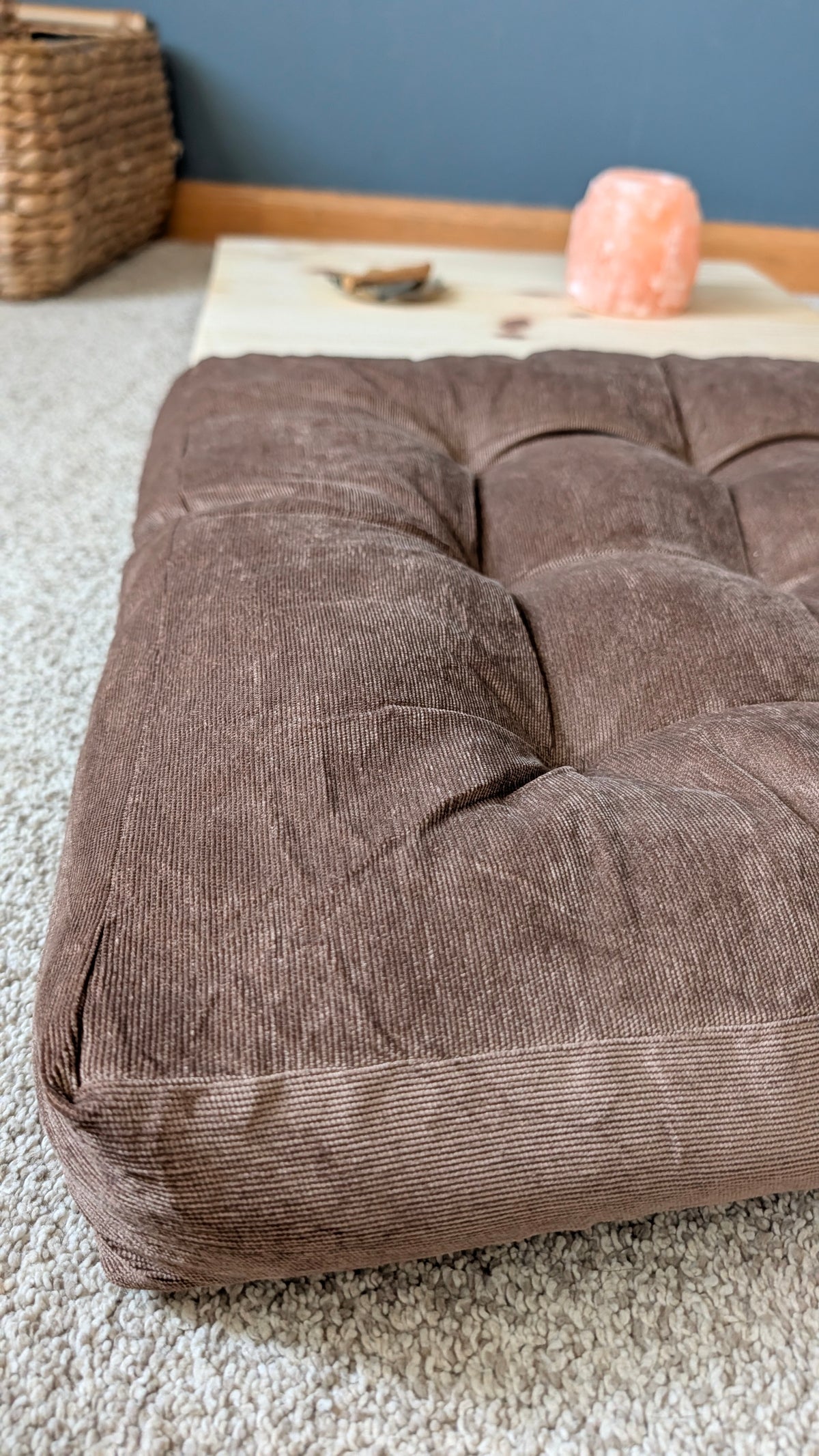 A large, tufted brown corduroy floor cushion on a light beige carpeted floor, with a blue wall and home decor items in the background.