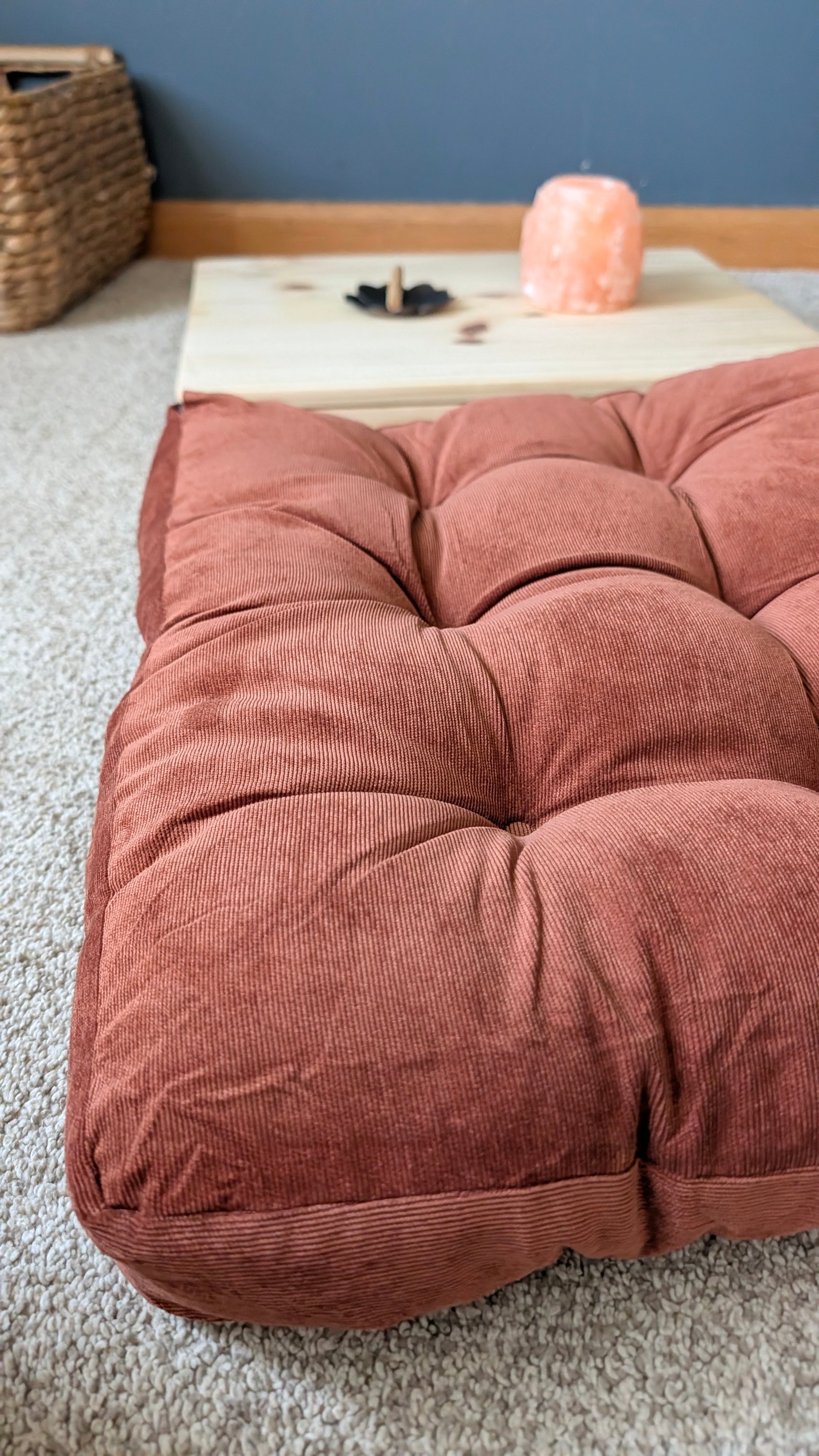A large, tufted floor cushion in a dusty terracotta rust color made of ribbed corduroy fabric, set on a light beige carpet in a room with a blue wall and a wooden table in the background.