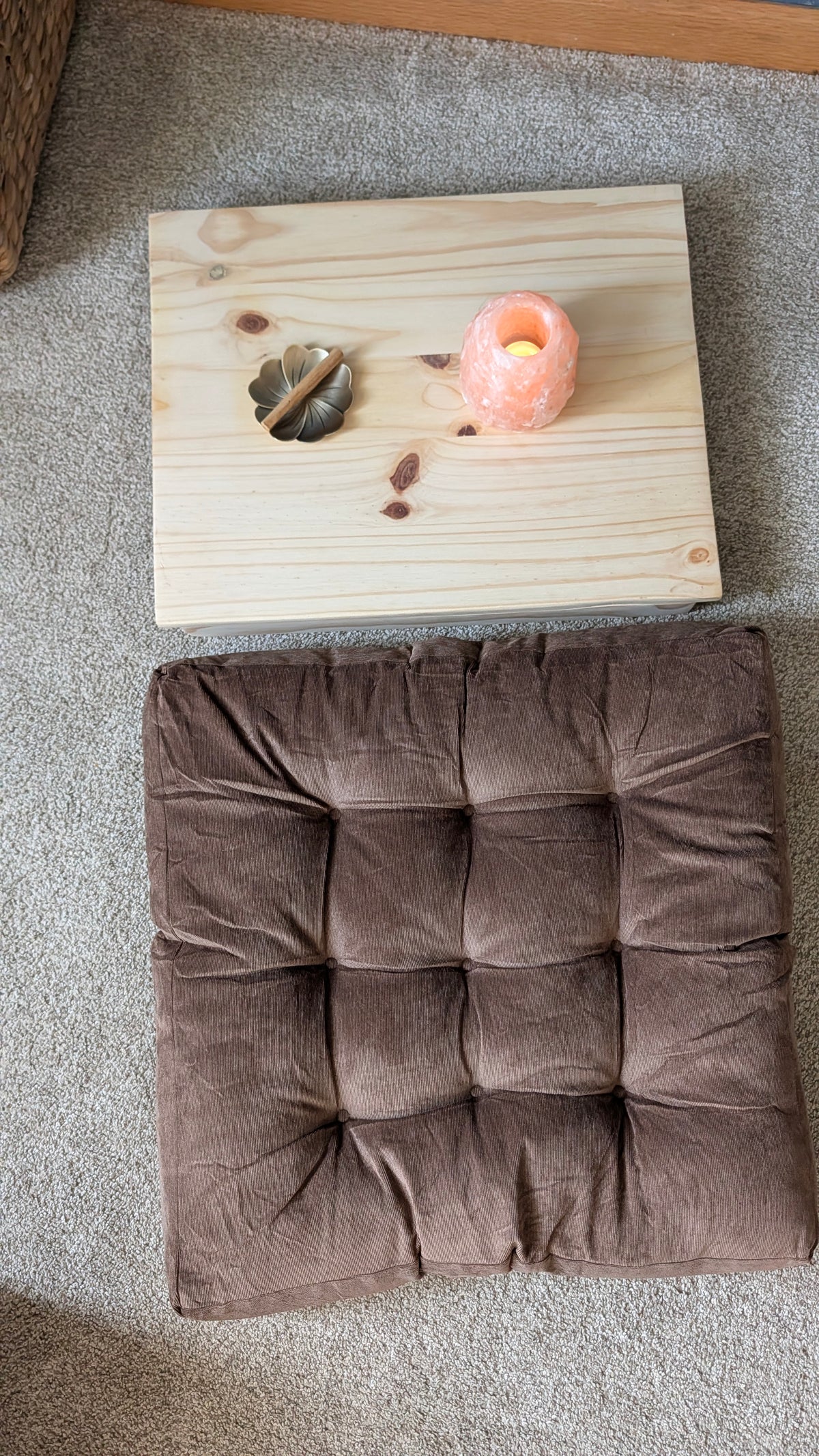 A square brown tufted cushion with a velvet-like texture, laid flat on a gray carpet next to a small wooden board holding a pink candle and a dried leaf.
