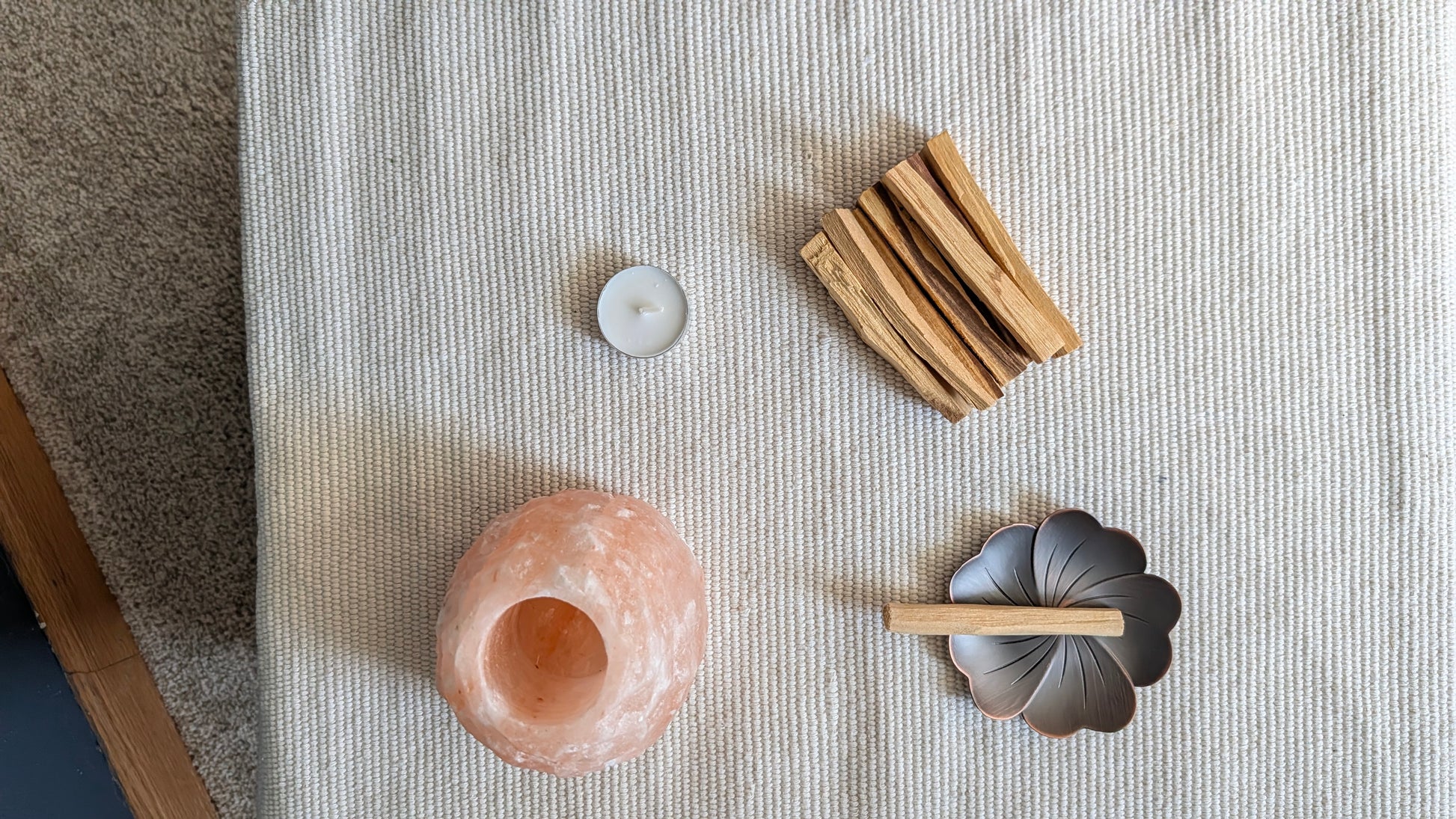 A styled flat-lay of wellness and spa accessories on a textured cream fabric surface, including a pink salt candle holder, a small tealight candle, a bundle of wooden sticks, and a dark metal flower-shaped holder with a wooden stick.