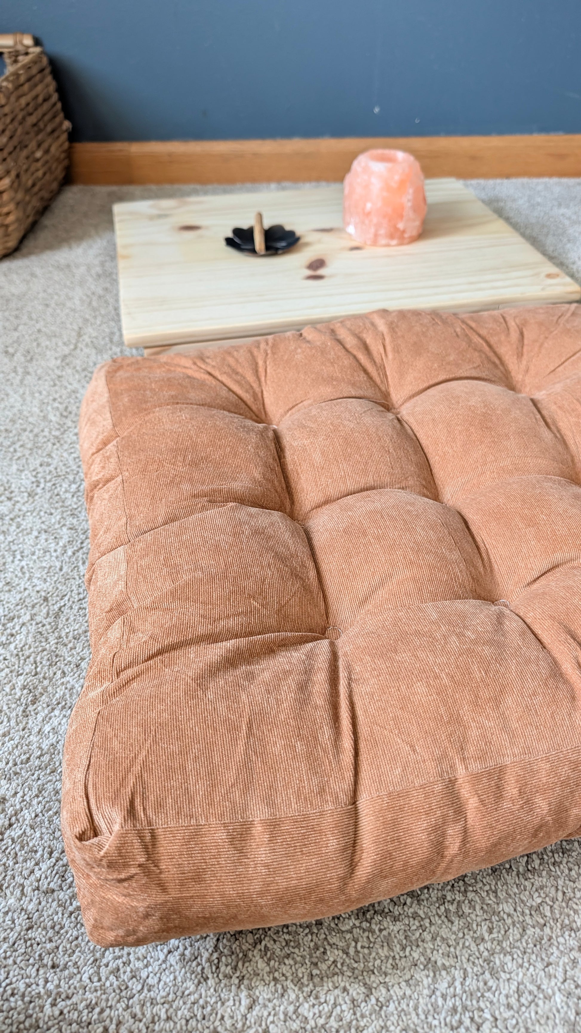 A large, tufted floor cushion in a warm terracotta or rust color, styled on a light beige carpet with a small wooden tray and a pink crystal salt lamp in the background.