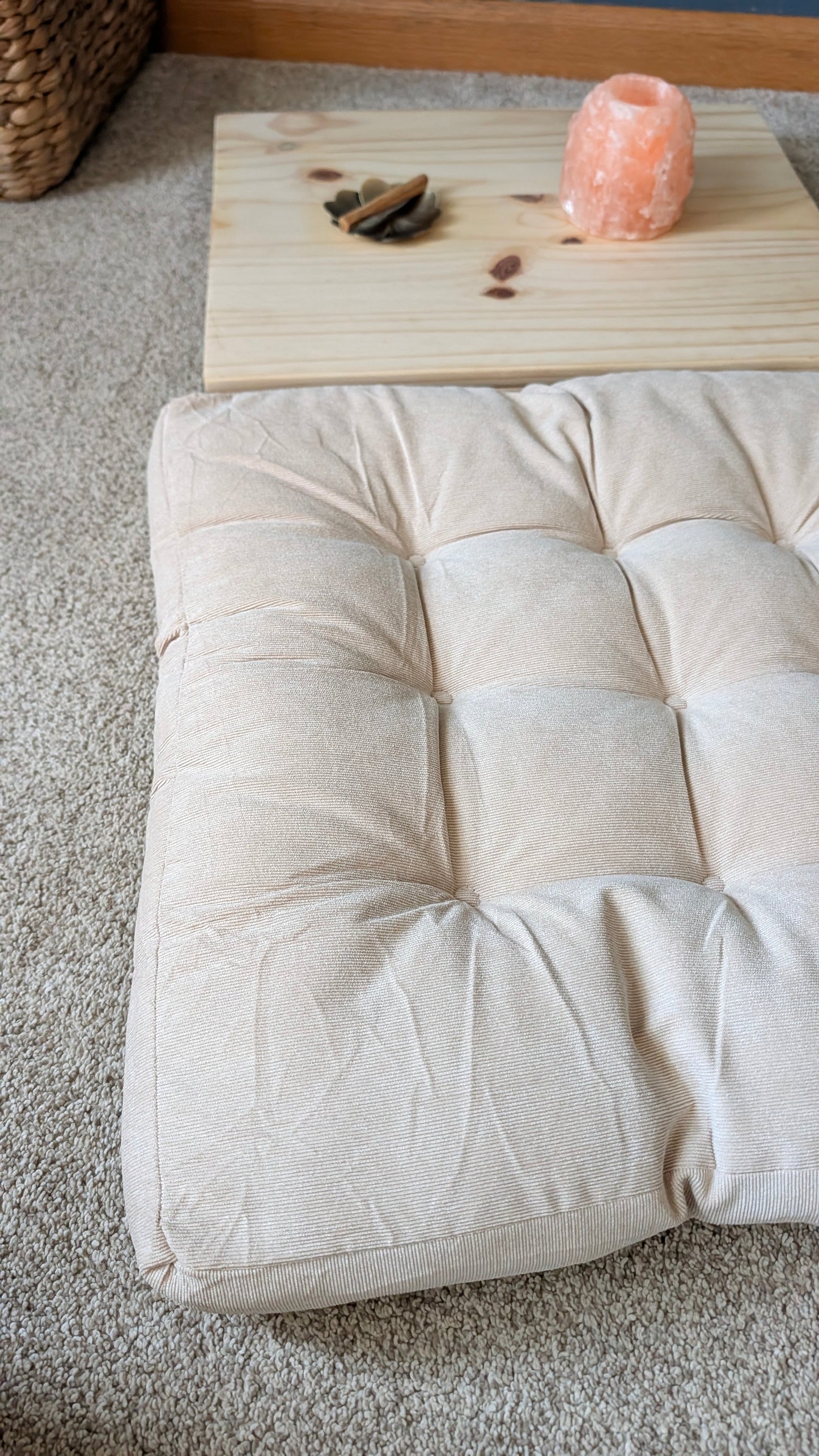A square tufted floor cushion in a light beige fabric, styled on a gray carpet with a wooden tray and a pink salt lamp in the background.