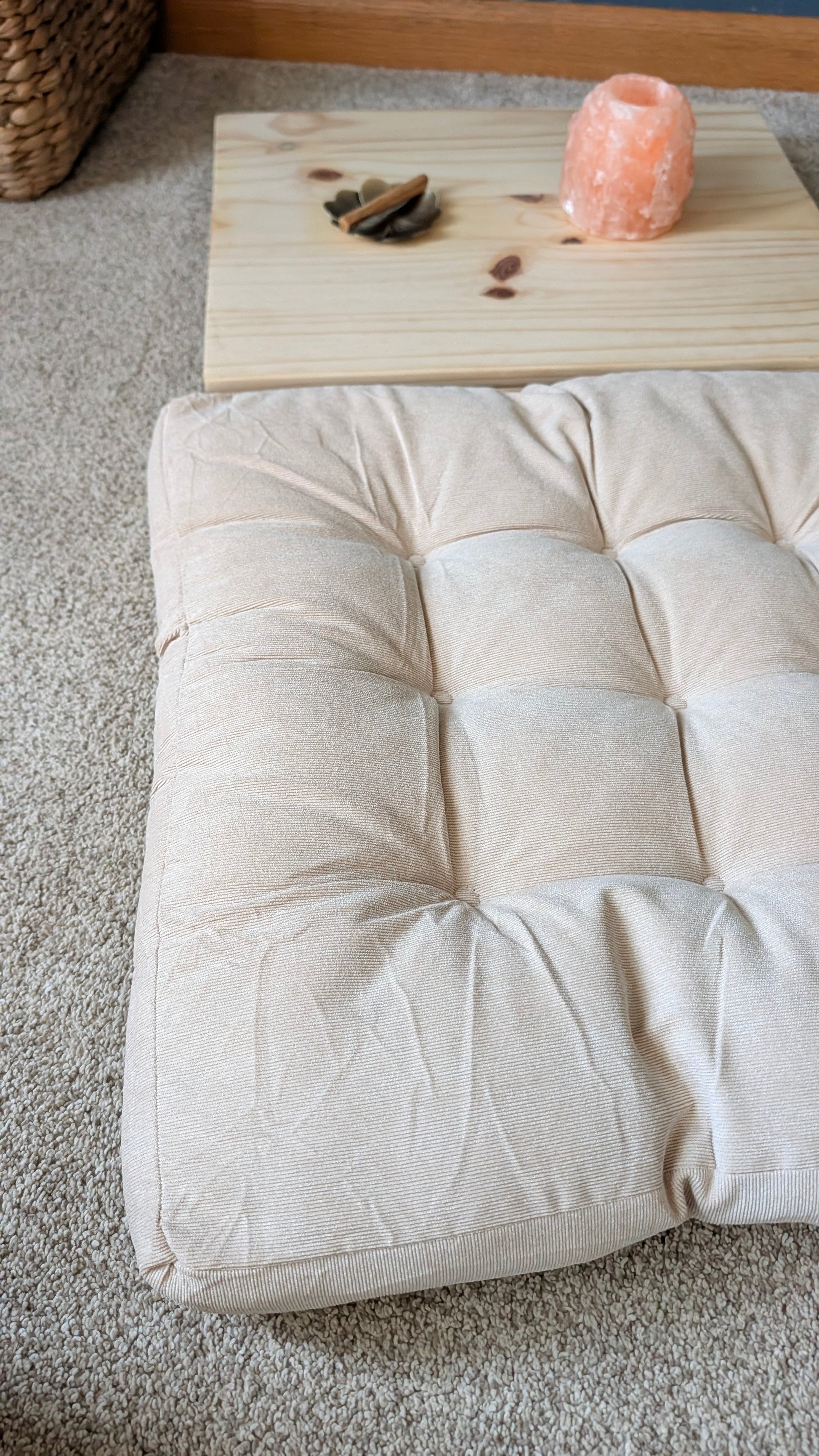 A square tufted floor cushion in a light beige fabric, styled on a gray carpet with a wooden tray and a pink salt lamp in the background.