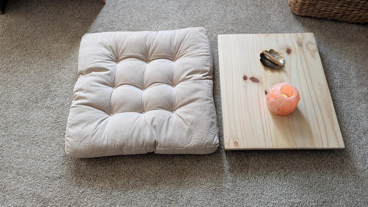 A square tufted floor cushion in a light beige fabric sits next to a small rectangular wooden tray on a gray carpet. The tray holds a small metallic object and a pink candle.
