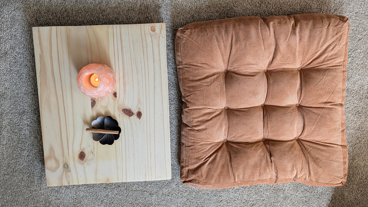A square tufted cushion in a warm camel-brown velvet fabric lies on a gray carpet next to a small wooden tray holding a lit himilayan salt tea light votive and a flower shaped metal tray holding palo santo.