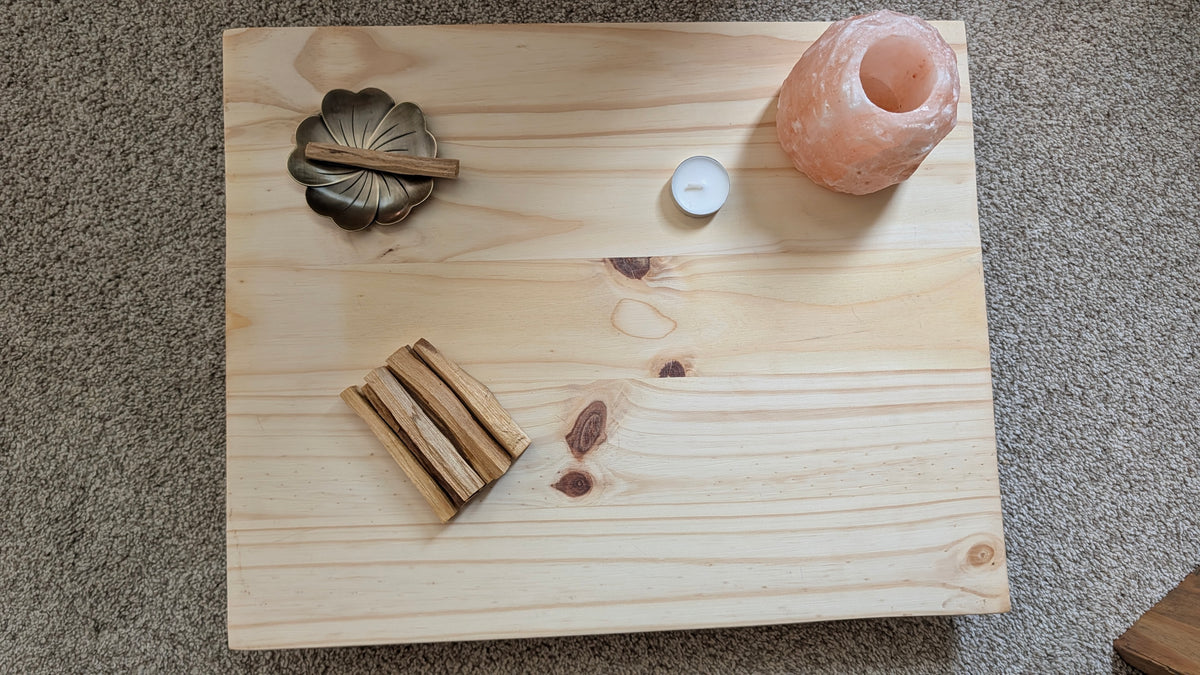 A square light wood tray or serving board placed on a beige carpet, styled with a small metal dish, cinnamon sticks, a white tea light candle, and a pink salt lamp.